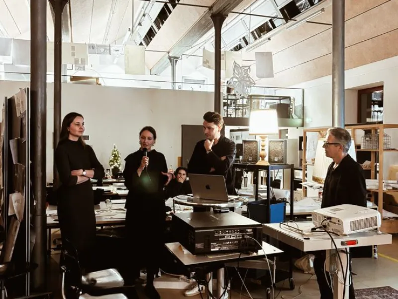 Eine Gruppe von Personen steht in einem modernen Architekturstudio. Eine Person spricht in ein Mikrofon, während andere aufmerksam zuhören. Im Raum sind technische Geräte und Präsentationsmaterialien zu sehen.