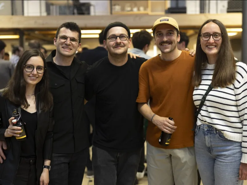 Eine Gruppe von fünf Personen lächelt in die Kamera bei einem Indoor-Event. Zwei Personen halten Getränke in der Hand. Die Umgebung ist lebhaft und gut beleuchtet, was auf eine gesellige Atmosphäre hinweist.