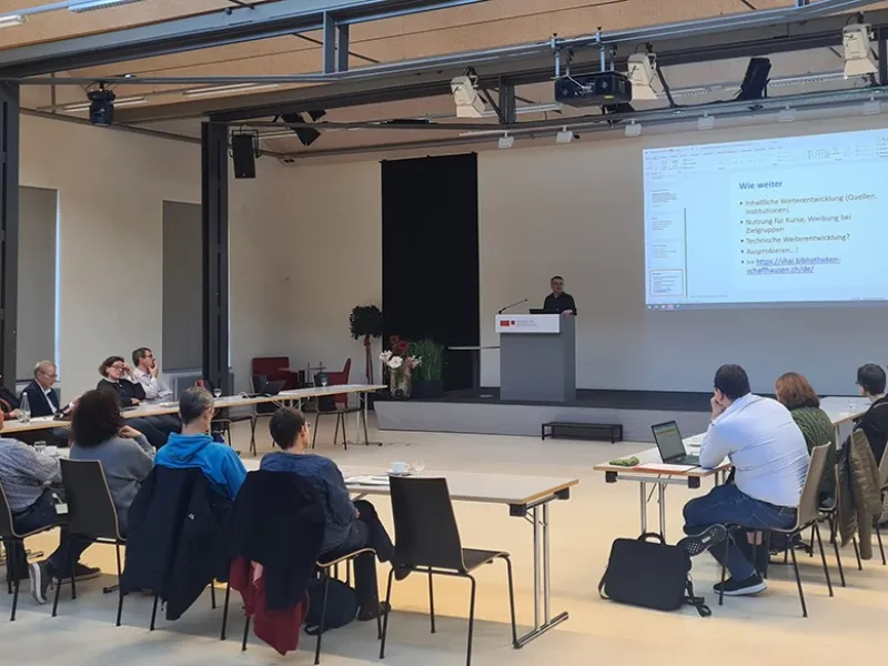 Eine Gruppe von etwa 20 Personen sitzt in einem Auditorium der Universität Liechtenstein, während ein Vortragender an einem Rednerpult spricht. Auf einer großen Leinwand ist eine Präsentation mit dem Titel "Wie weiter" zu sehen.